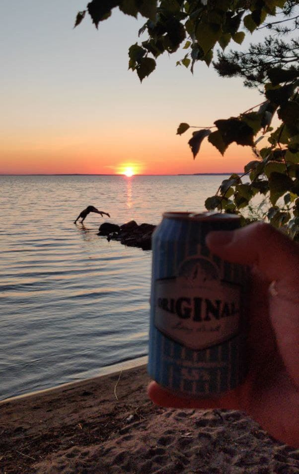 My hand holding a long drink in, with a person jumping into water in the background and a beautiful sunset.